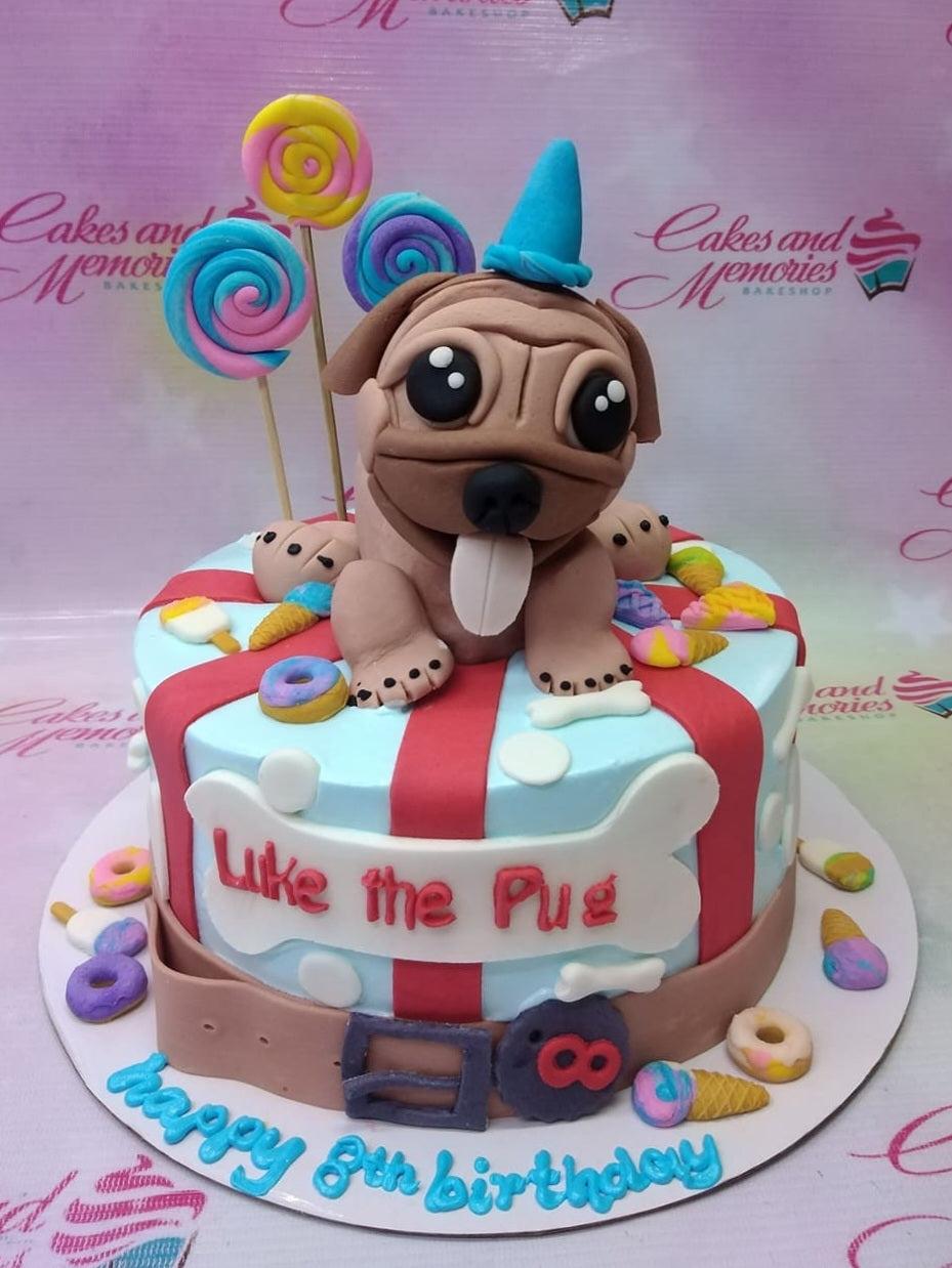 Pug themed birthday cake with a large fondant pug figure, colorful lollipops, donuts, and ice cream decorations on a blue and red striped base.