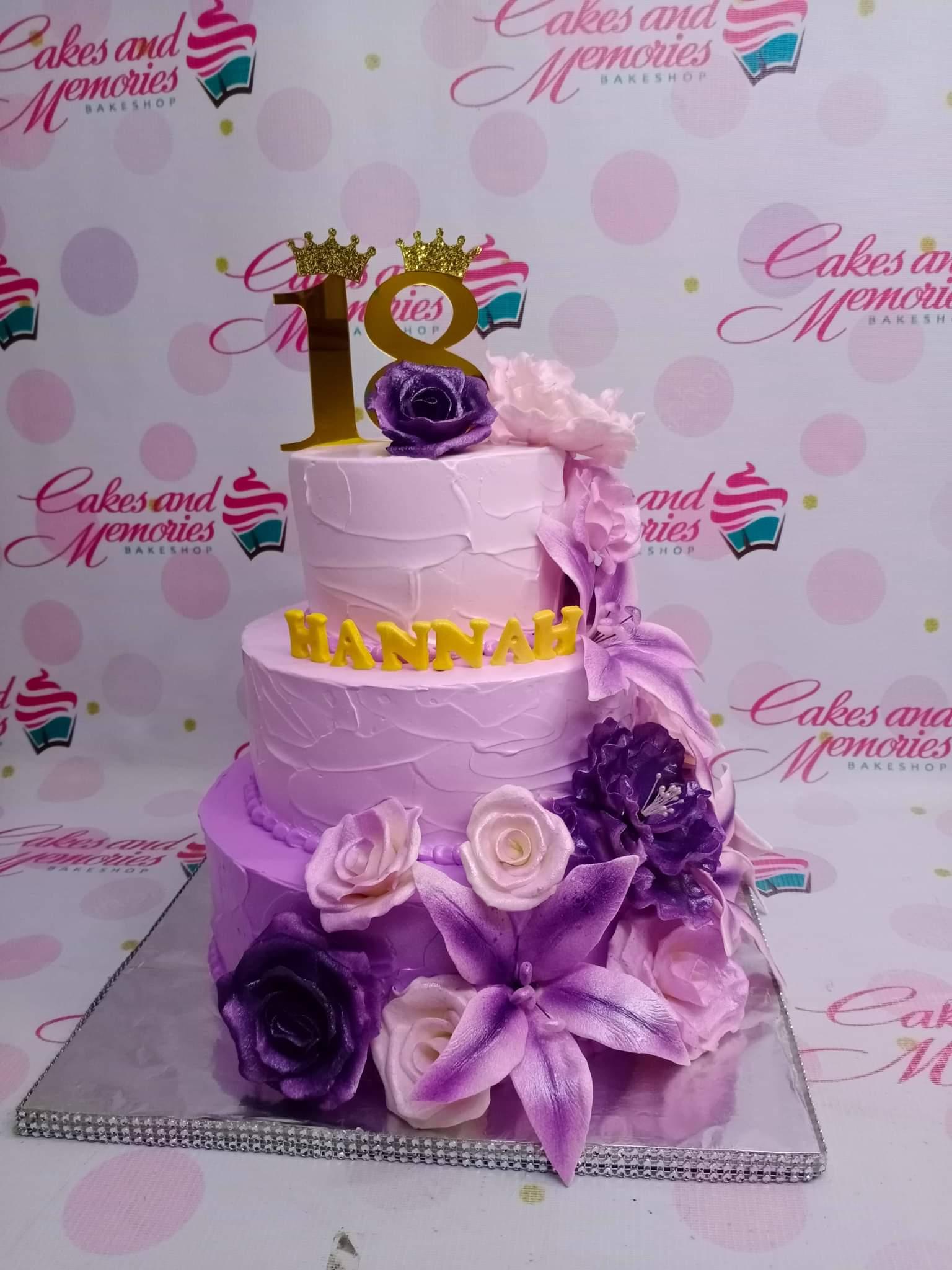 Three-tier purple and pink ombre cake with large gumpaste flowers and a gold number 18 topper.