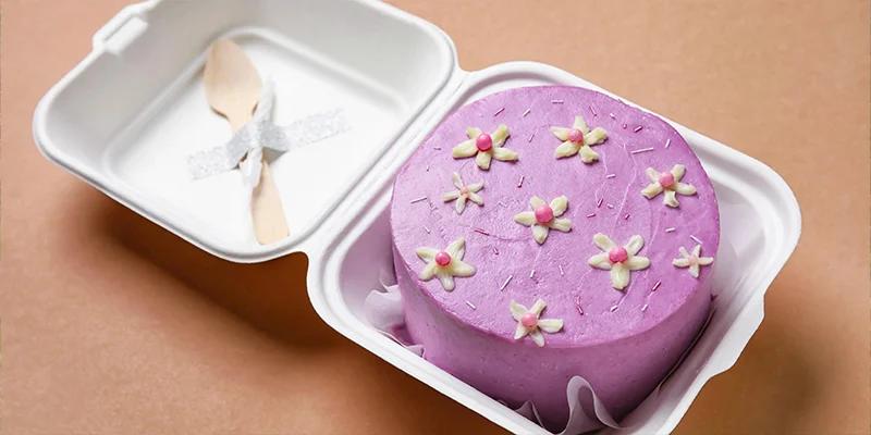 Purple bento cake with white piped icing flowers and pink sugar pearls in a white clamshell box with a wooden spoon.