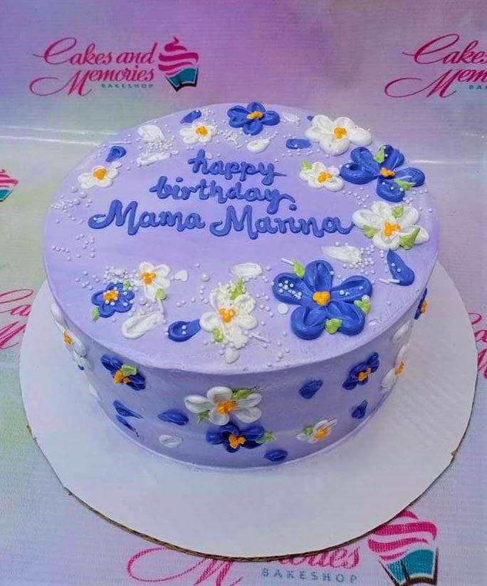 Purple round cake decorated with blue and white piped icing flowers and white sprinkles for a birthday celebration.