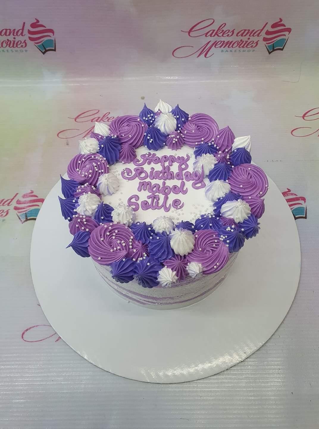 Purple and white floral-themed birthday cake with intricate icing rosettes and white pearl sprinkles.