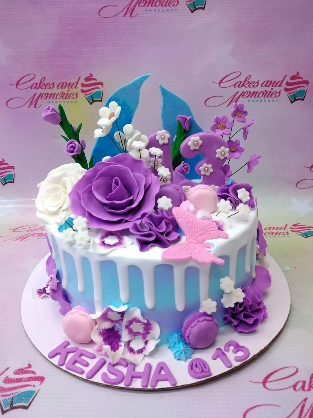 Purple and blue ombre cake with white icing drip, decorated with large purple fondant roses, pink butterflies, and macarons.