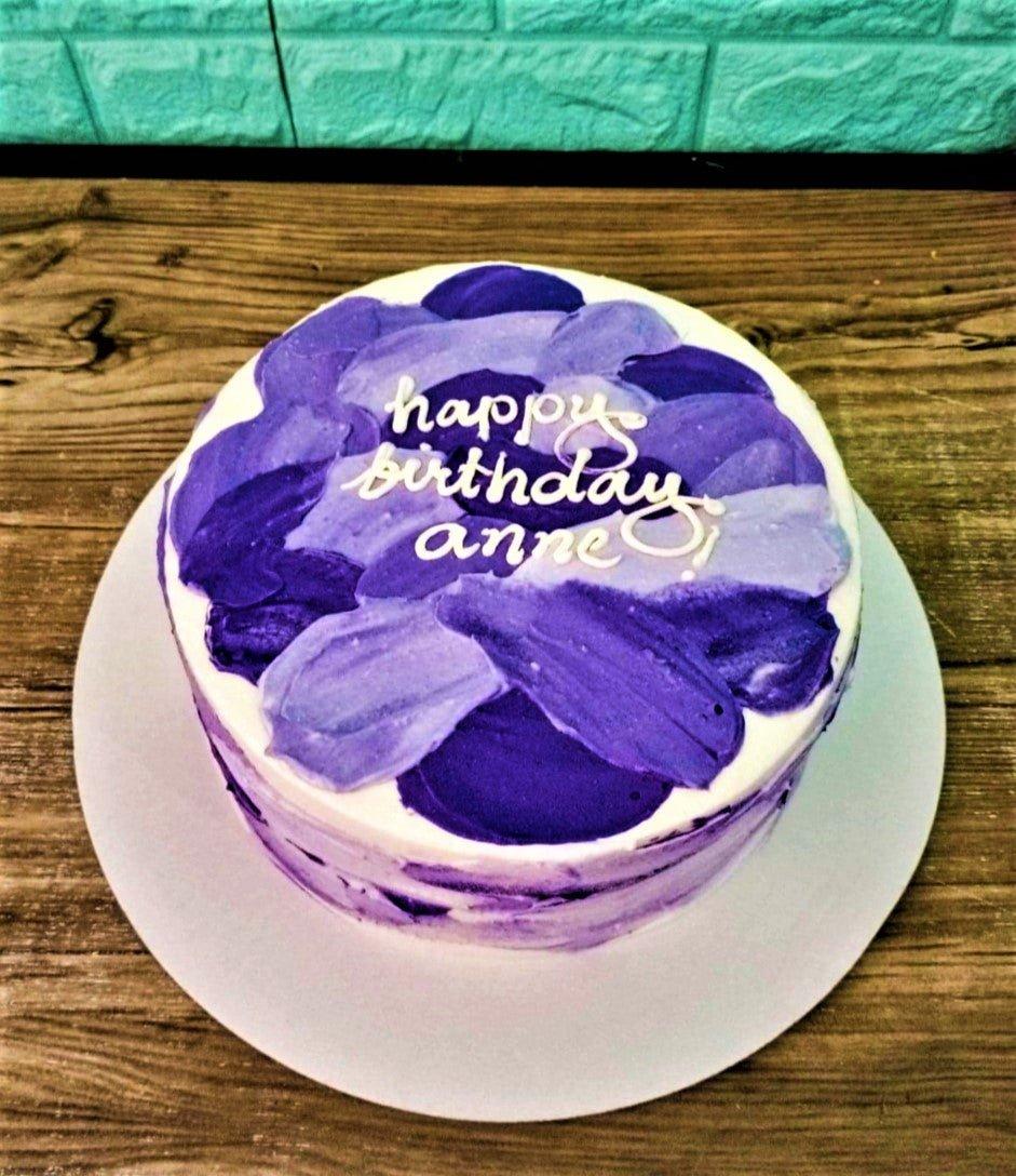 Minimalist white cake with purple and lavender palette knife icing strokes and a white piped Happy Birthday Anne message.