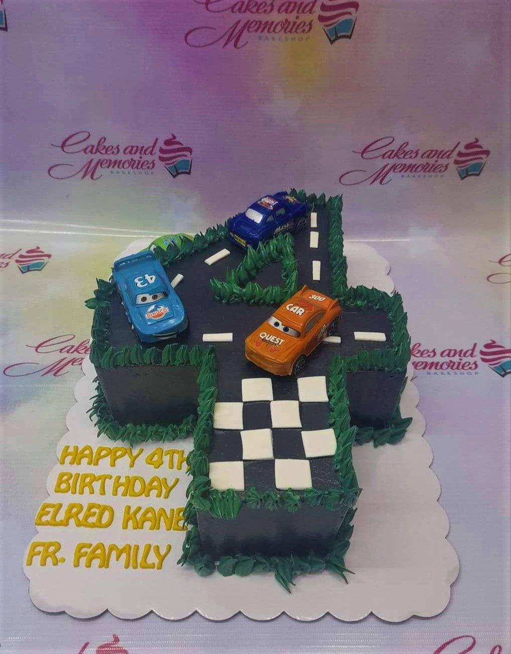 Number 4 shaped racing car birthday cake with blue, orange, and navy toy cars on a black track with green icing grass.