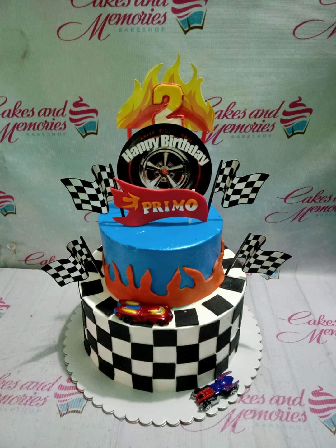 Two-tier racing themed birthday cake with checkered pattern, blue top tier, orange flames, and toy cars.