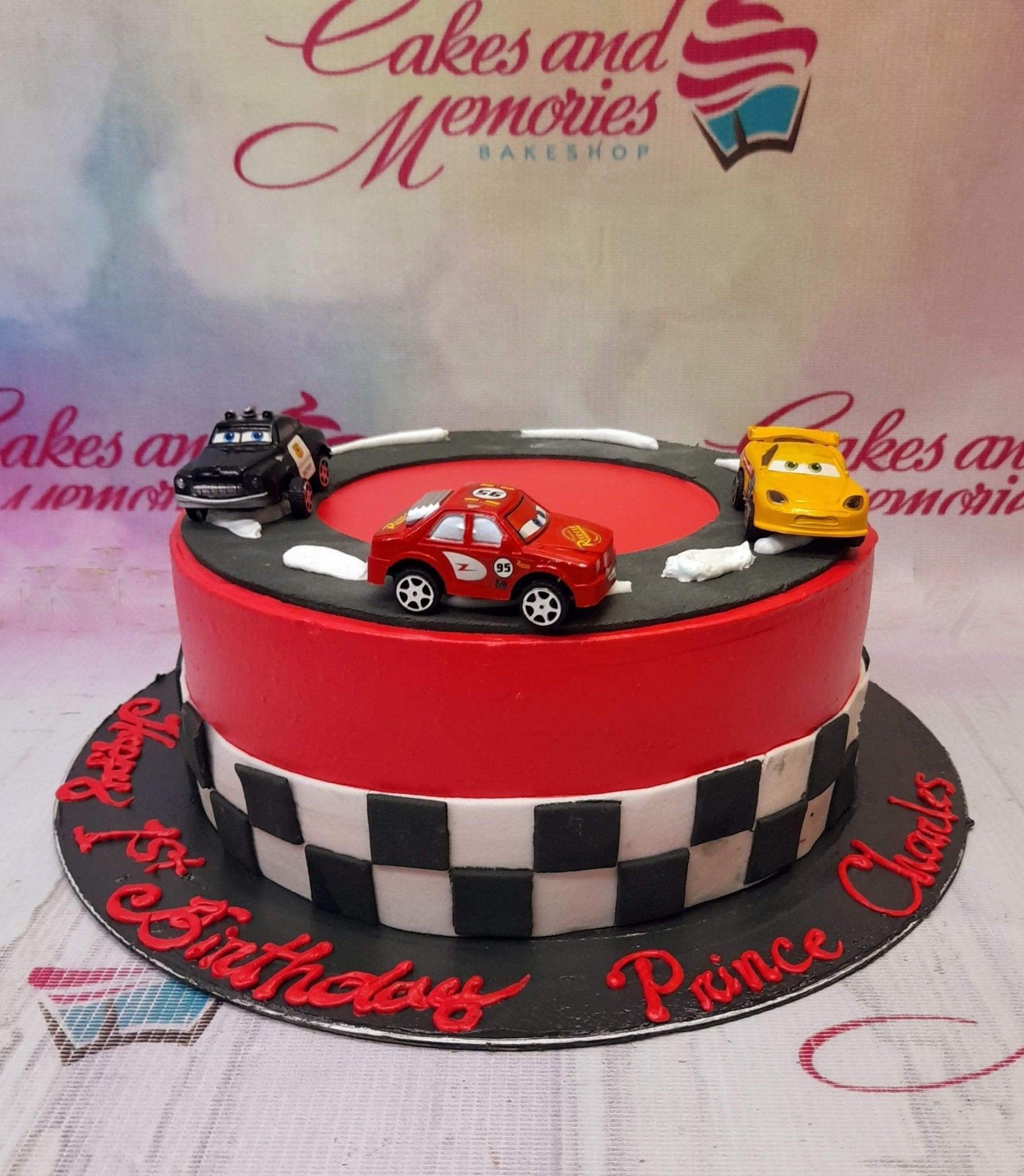 Red and black racing car themed birthday cake with toy cars and checkered flag pattern on the side.