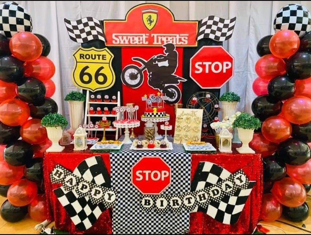 Red and black racing themed birthday cake with checkered patterns and motorcycle toppers on a dessert table.