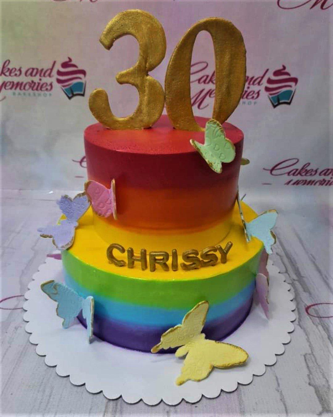 A vibrant two-tier rainbow gradient cake with gold number 30 topper, colorful butterflies, and the name Chrissy in gold letters.