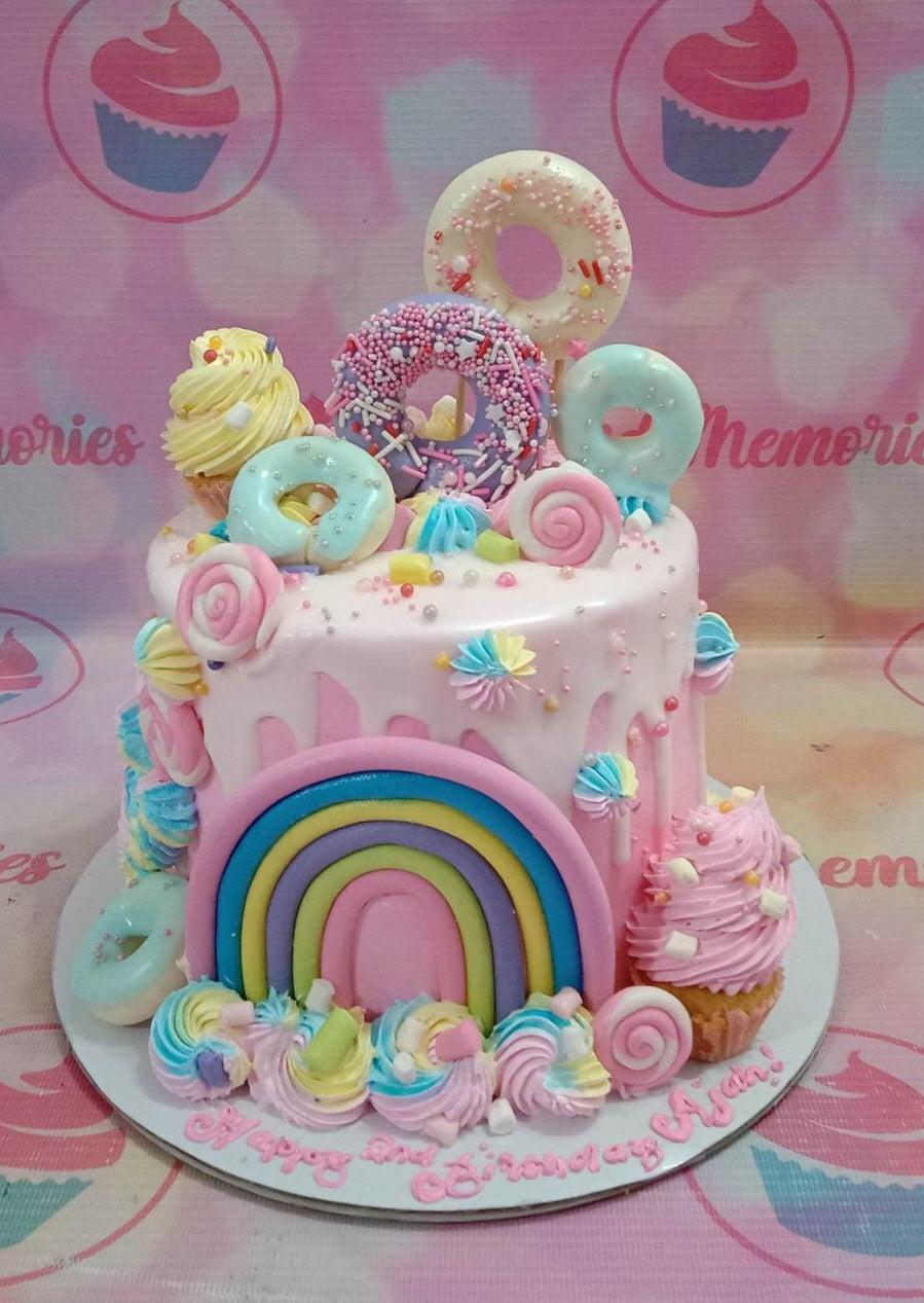 Pink drip cake with rainbow fondant, donuts, cupcakes, and colorful icing swirls for a 2nd birthday celebration.