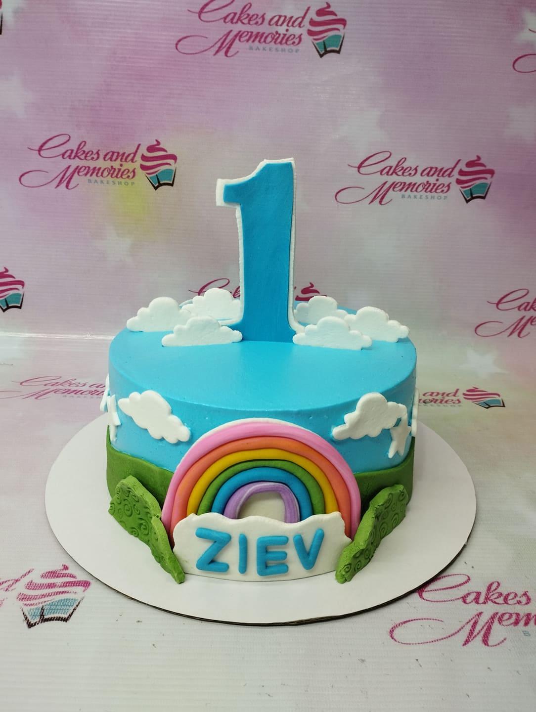 Light blue 1-tier birthday cake with a large blue number one topper, fondant rainbow, white clouds, and green hills with the name Ziev.