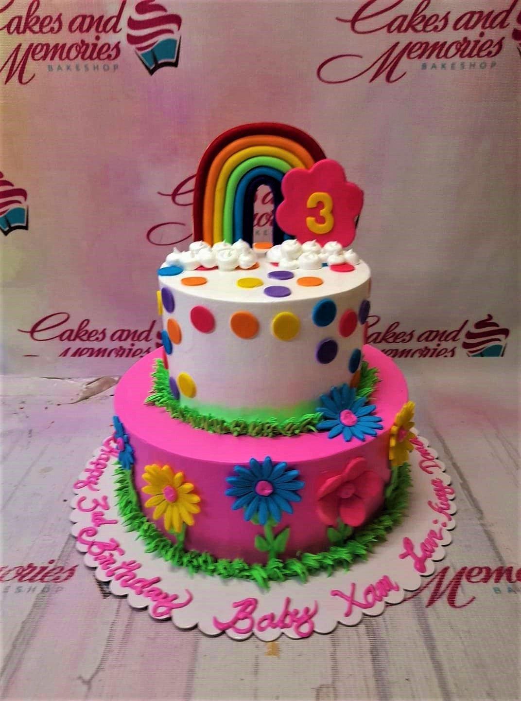 Two-tier rainbow and flower birthday cake with pink and white icing, colorful polka dots, and a large rainbow topper.
