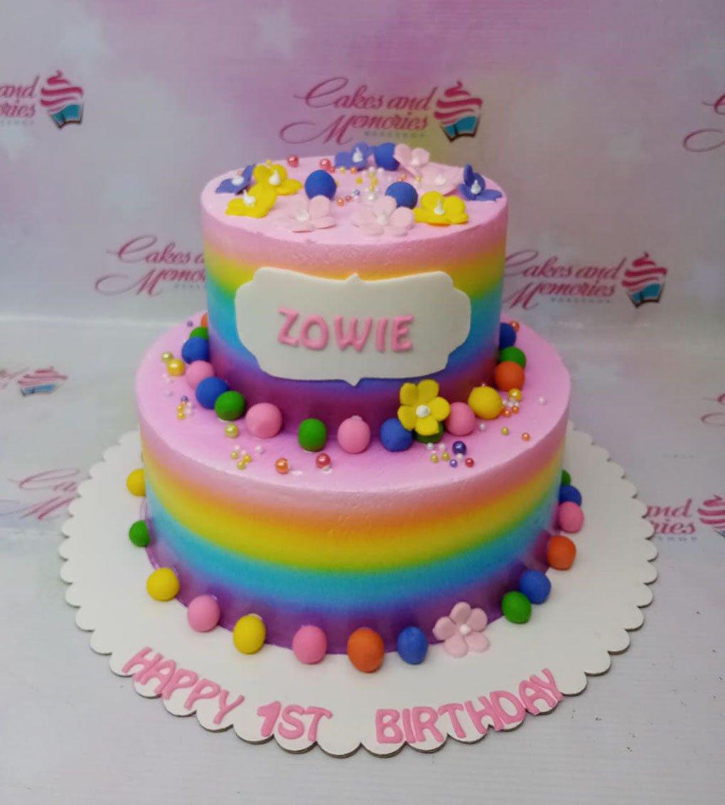 Two-tier rainbow gradient birthday cake with colorful fondant balls and flowers for Zowie's 1st birthday.