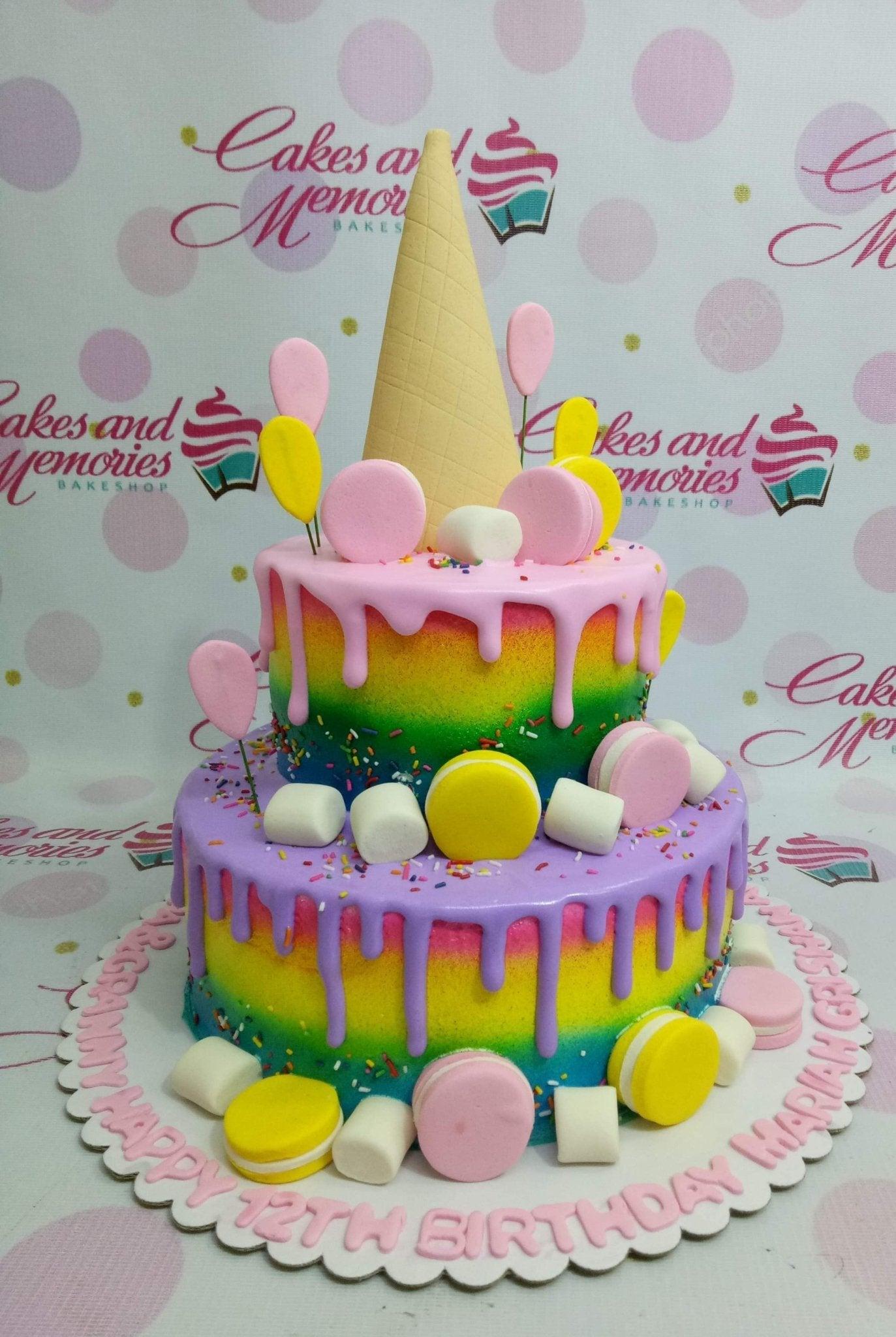 Two-tier rainbow drip cake with an ice cream cone topper, macarons, marshmallows, and colorful sprinkles.