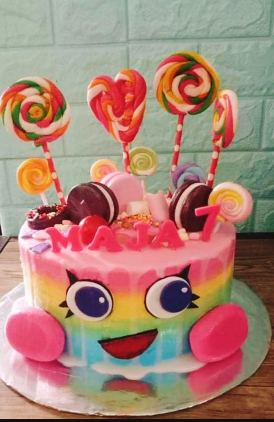 Rainbow themed birthday cake with colorful lollipops, macarons, and a cute character face on pink icing.
