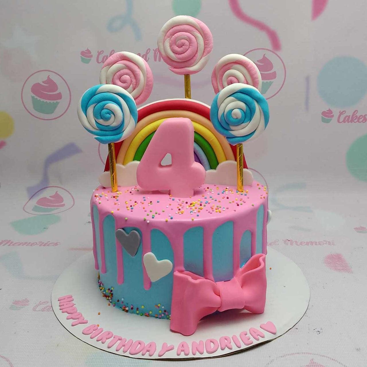 Light blue birthday cake with pink drip, rainbow topper, and colorful fondant lollipops for a 4th birthday celebration.