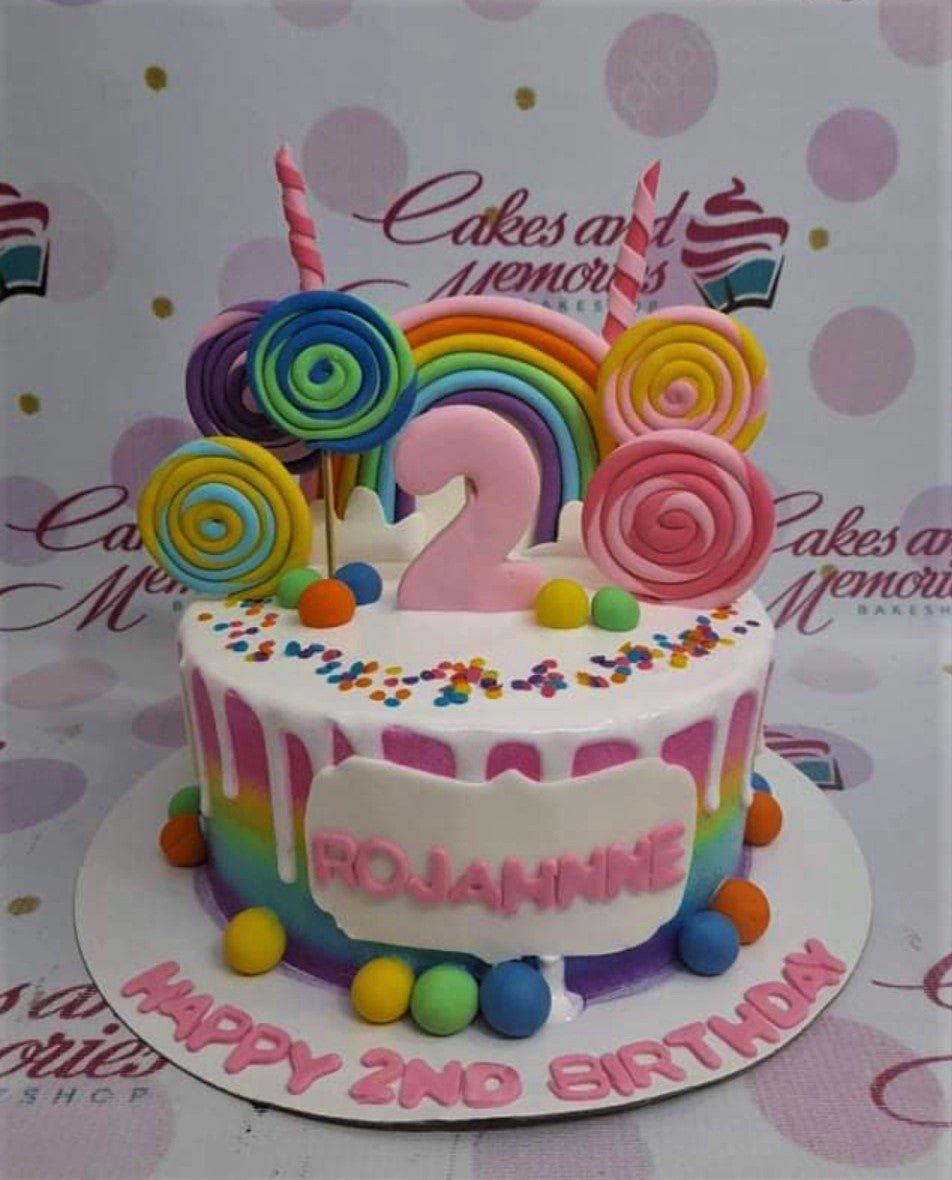 Rainbow themed 2nd birthday cake with colorful fondant lollipops, a large pink number 2, and white chocolate drip over a rainbow gradient base.