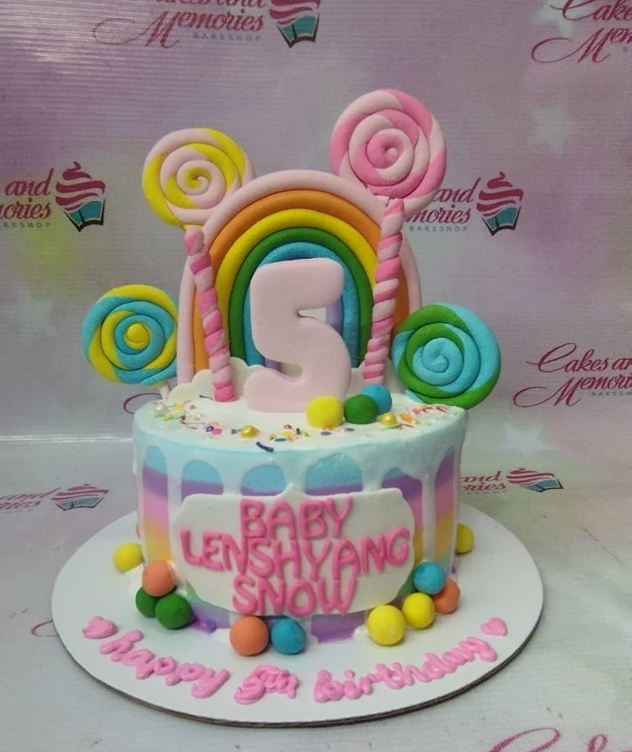 Colorful rainbow birthday cake with lollipop toppers, a large number 5, and a rainbow arch on a white base board.