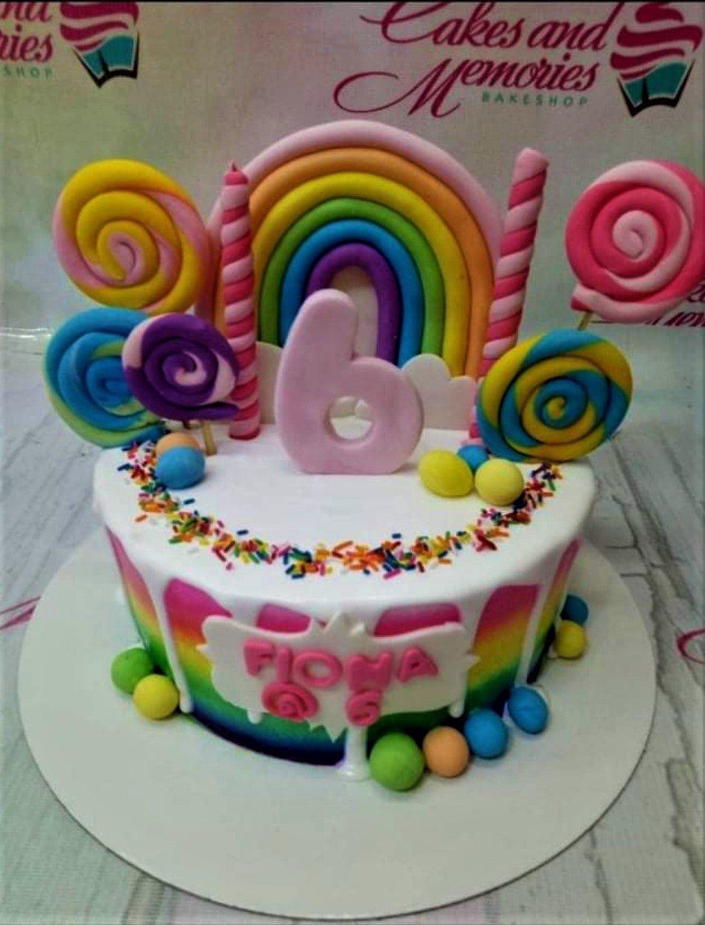 Rainbow themed birthday cake with colorful fondant lollipops, a large pink number 6, and a rainbow topper on white icing with sprinkles.