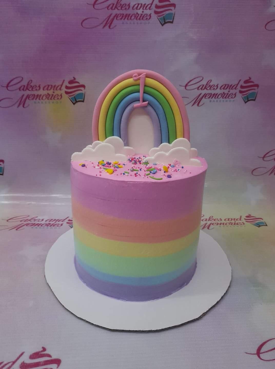 A tall single-tier rainbow birthday cake with a pastel rainbow topper, white fondant clouds, and colorful sprinkles on pink icing.