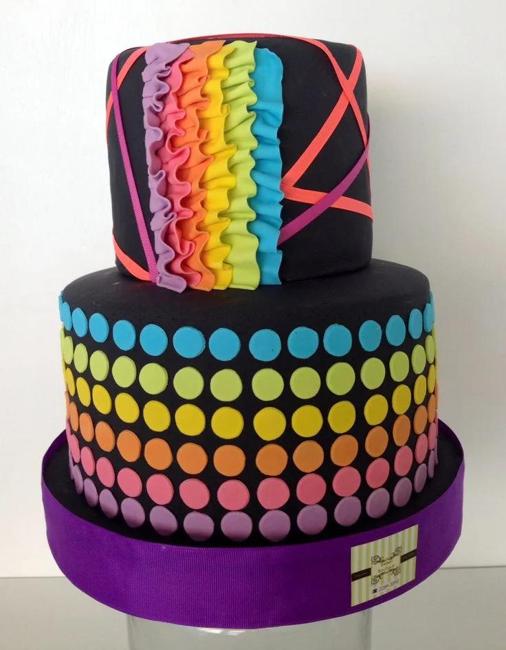Two-tier black fondant cake with vibrant rainbow ruffles and a colorful polka dot pattern on the bottom tier.