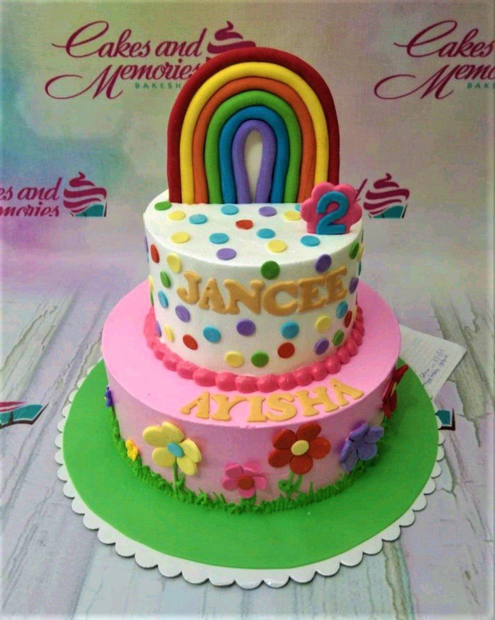 Colorful 2-tier rainbow birthday cake with polka dots, fondant flowers, and a large rainbow topper for Jancee Ayisha's 2nd birthday.
