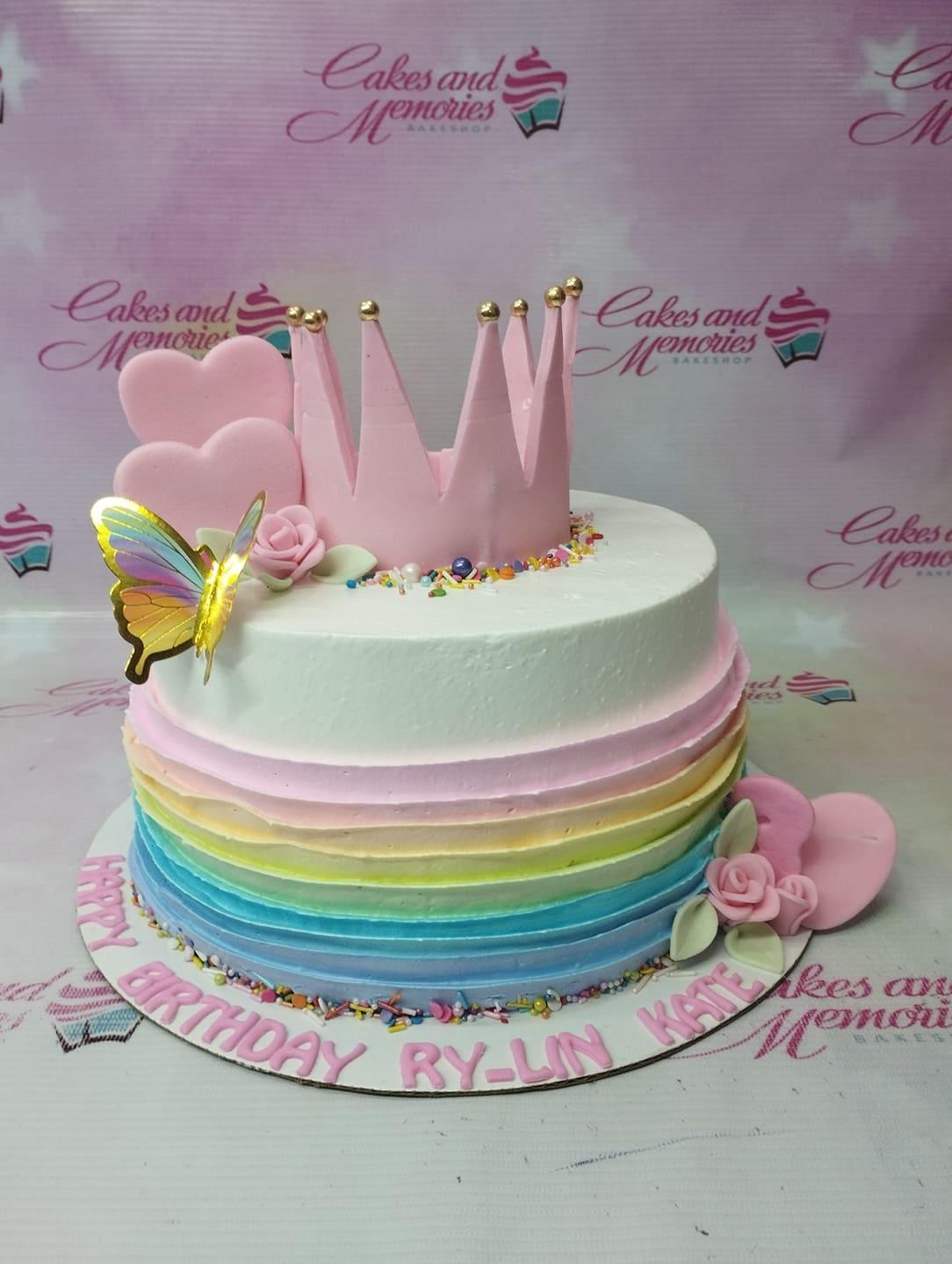 Single tier rainbow gradient cake with a pink fondant crown, pink hearts, and a colorful butterfly topper.