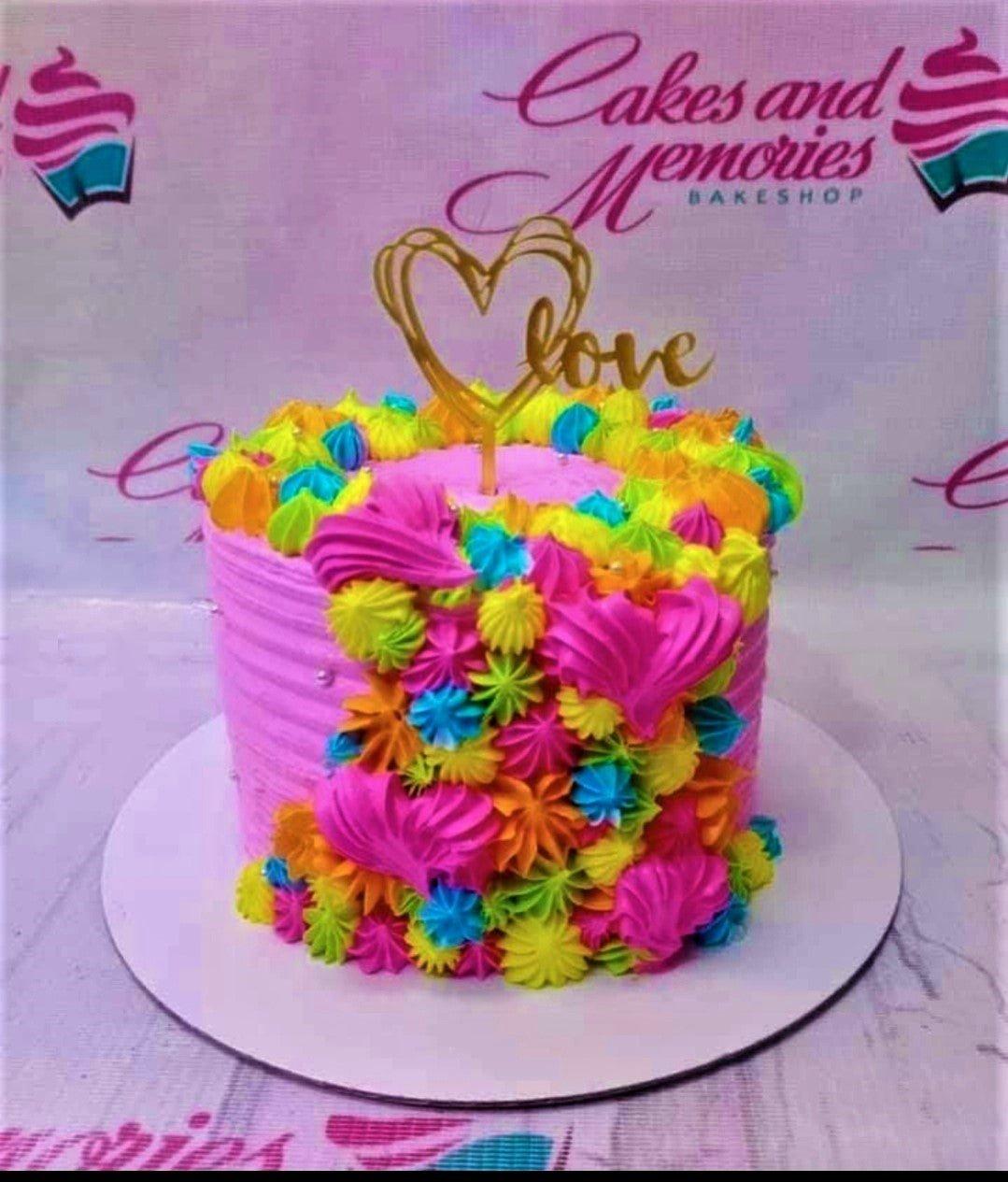 Vibrant pink cake with colorful rainbow icing rosettes and a gold heart love topper.