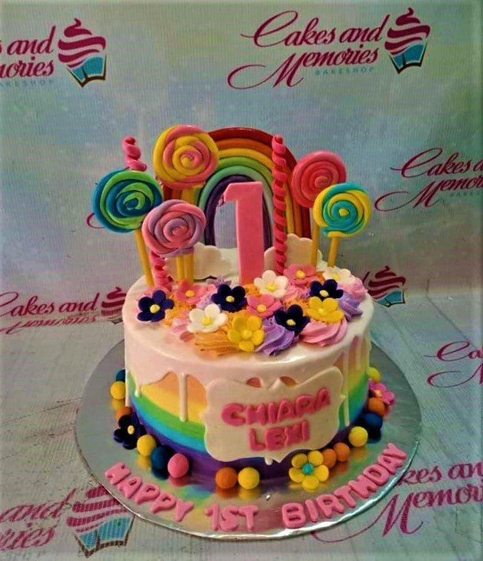 Rainbow themed 1st birthday cake with colorful fondant lollipops, a large pink number one, and a rainbow topper.