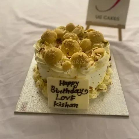 White soft-icing cake topped with round yellow sweets and crushed nuts, featuring a white chocolate message plaque.