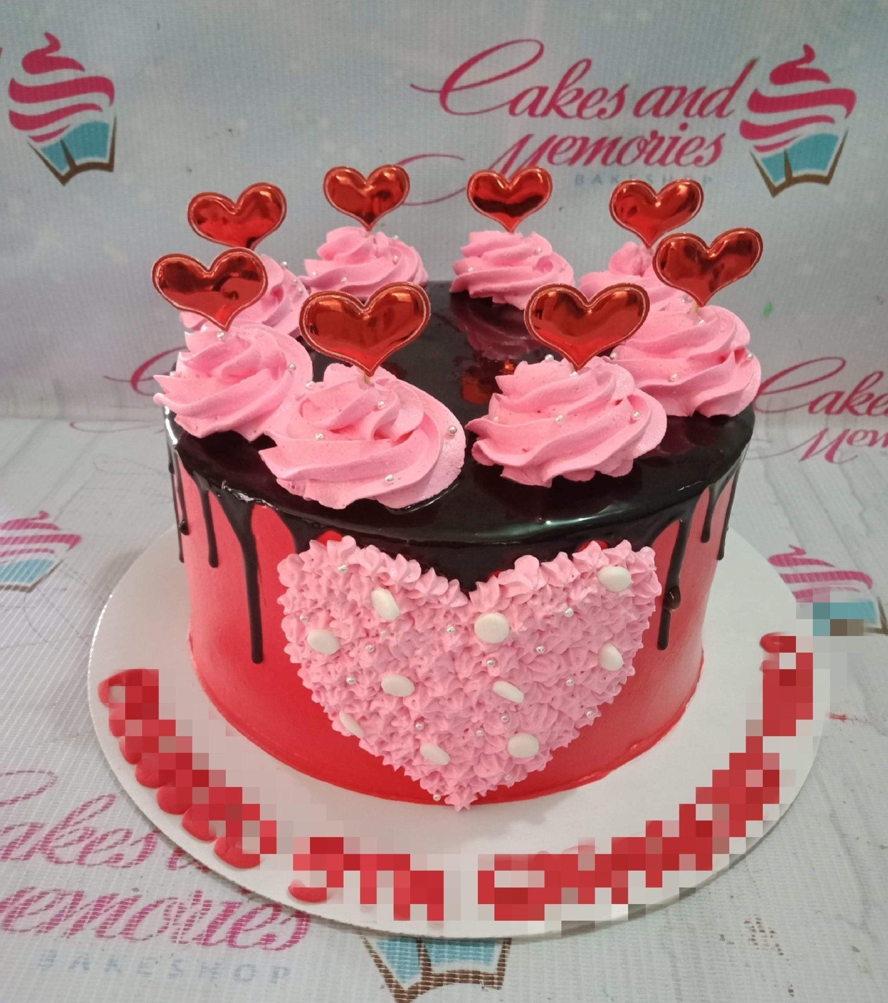 Red and pink heart-themed cake with chocolate drip and pink icing rosettes topped with red metallic heart toppers.
