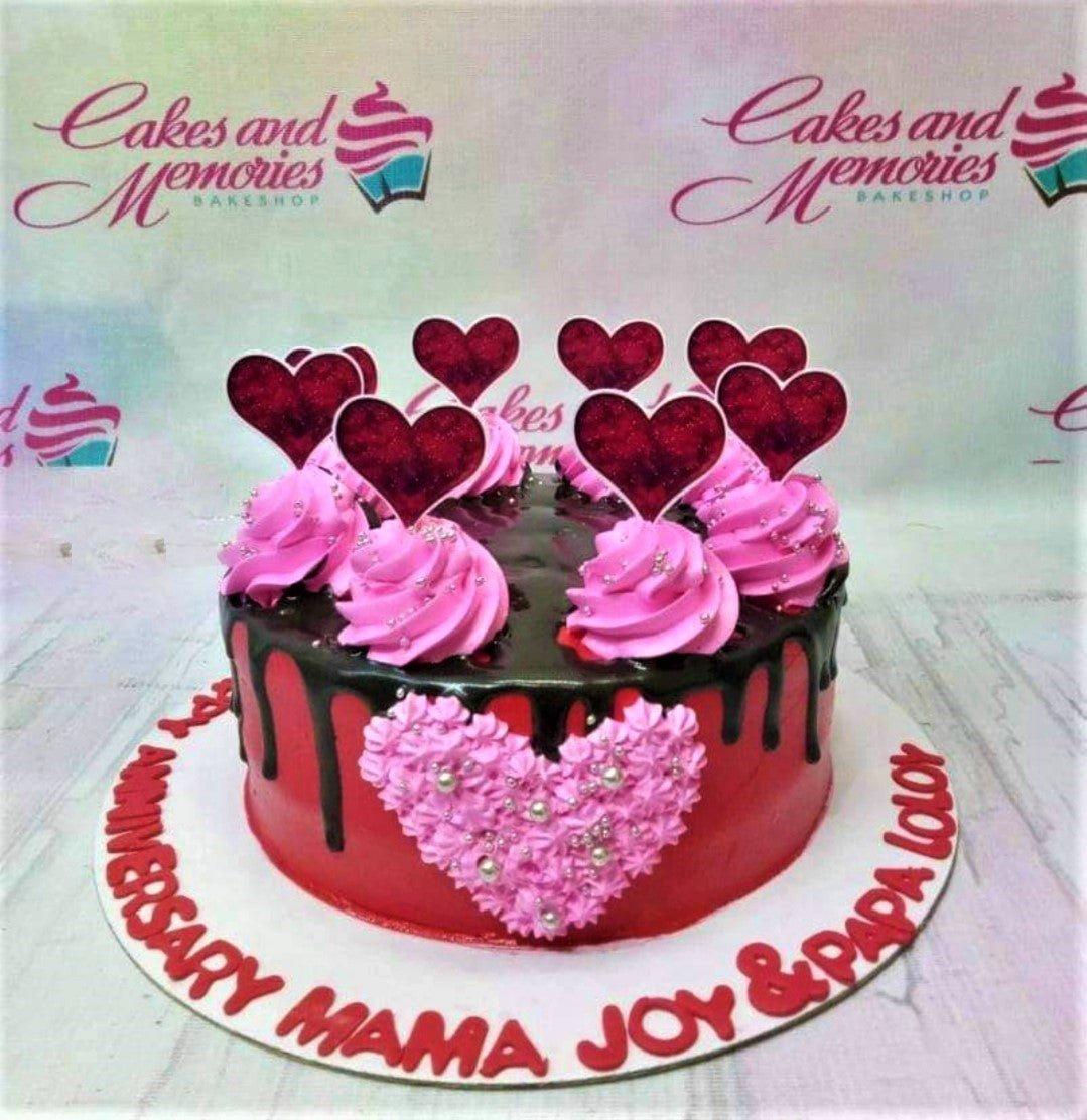 Red anniversary cake with chocolate drip, pink icing rosettes, and glittery red heart toppers.