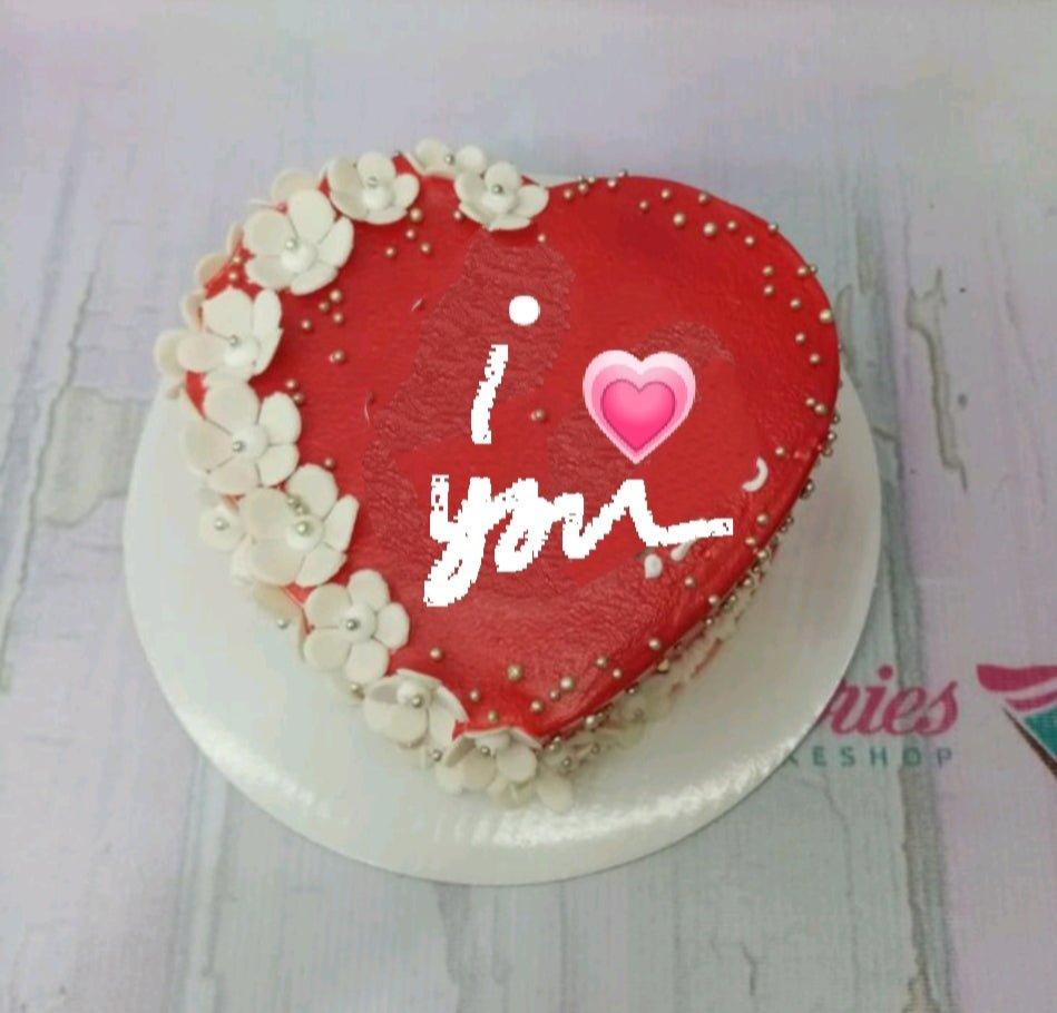 Heart-shaped red cake with white fondant flowers and silver dragees featuring an I Love You message.