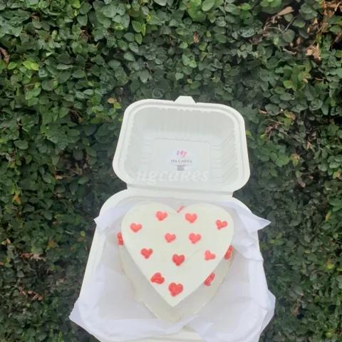 Heart-shaped white bento cake with small red icing hearts on top and sides, presented in a white clamshell box.