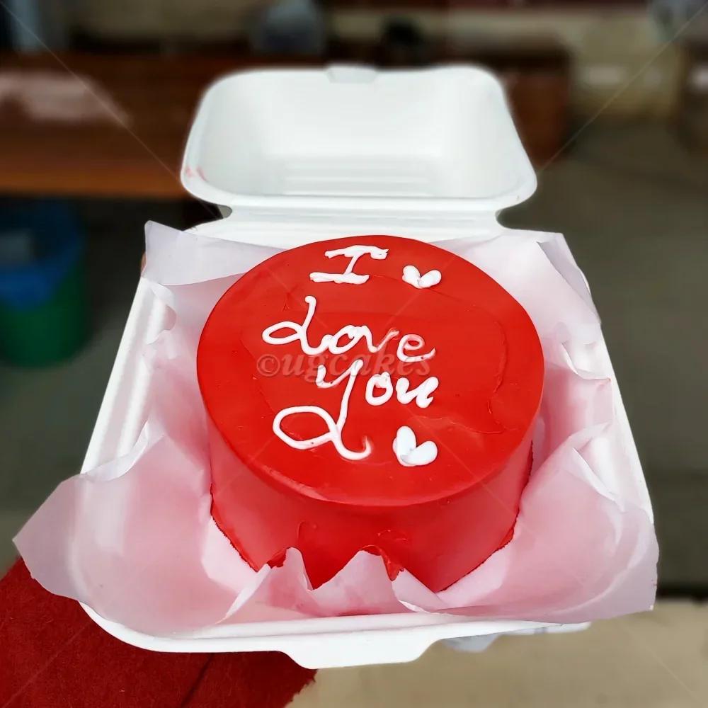 Red minimalist bento cake with white piped icing message I Love You and small heart accents in a white styrofoam box.