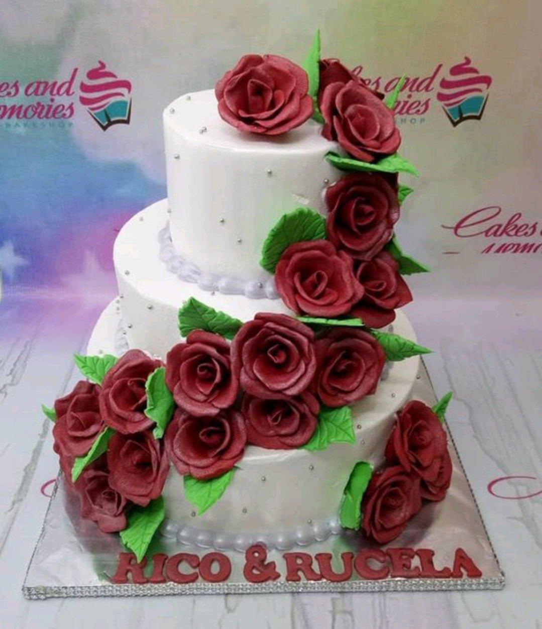Elegant 3-tier white wedding cake adorned with a cascading trail of deep red gumpaste roses and green leaves on a silver board.