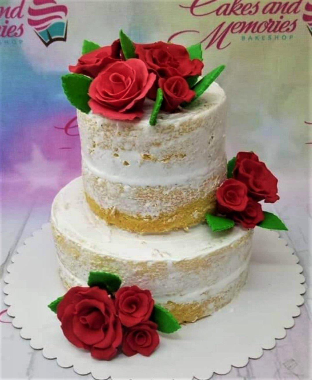 Two-tier semi-naked cake decorated with clusters of deep red gumpaste roses and green leaves on a white scalloped base.