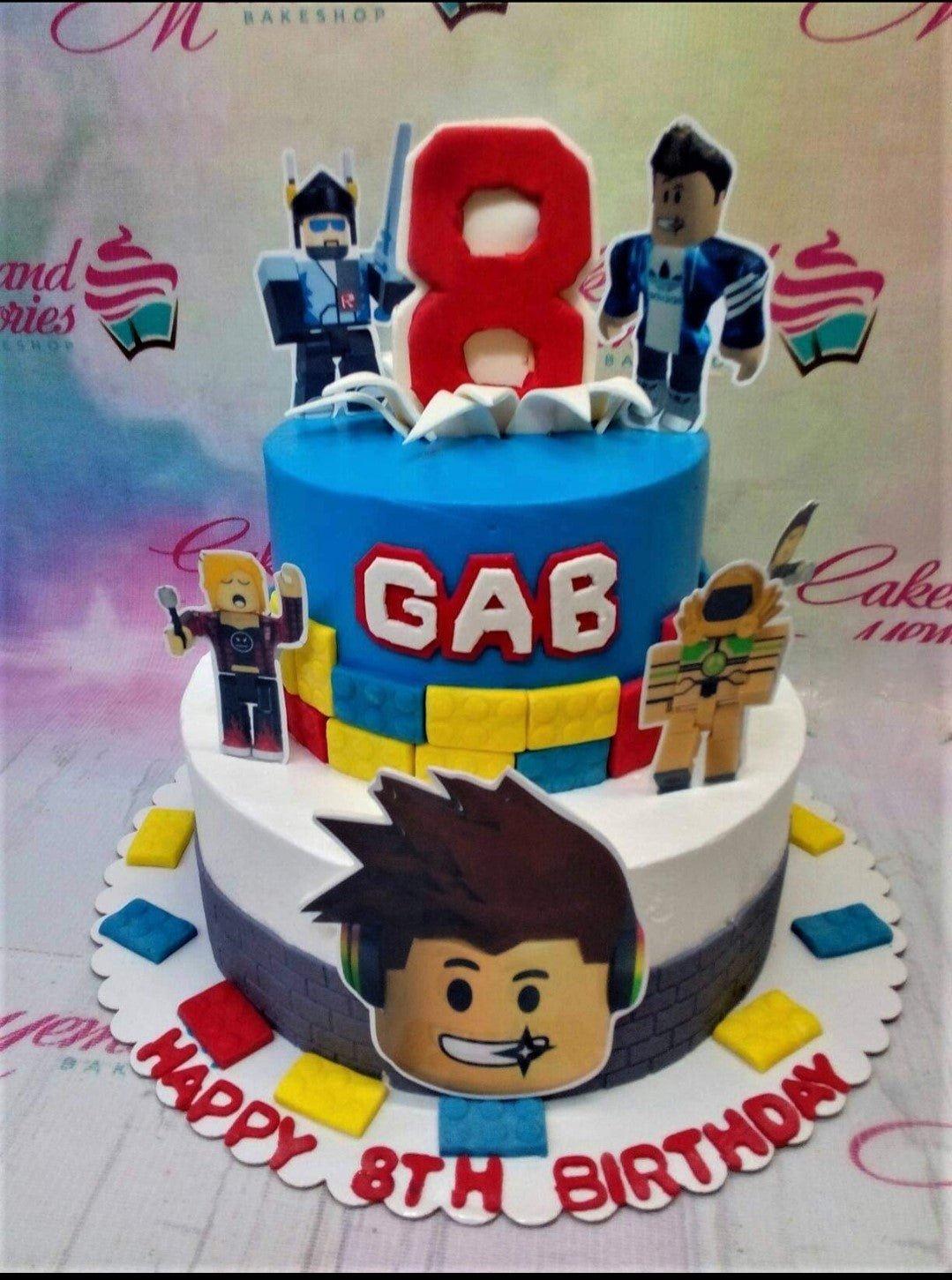 Two-tier Roblox themed birthday cake with blue and white icing, featuring character printouts, fondant building blocks, and a red number 8 topper.