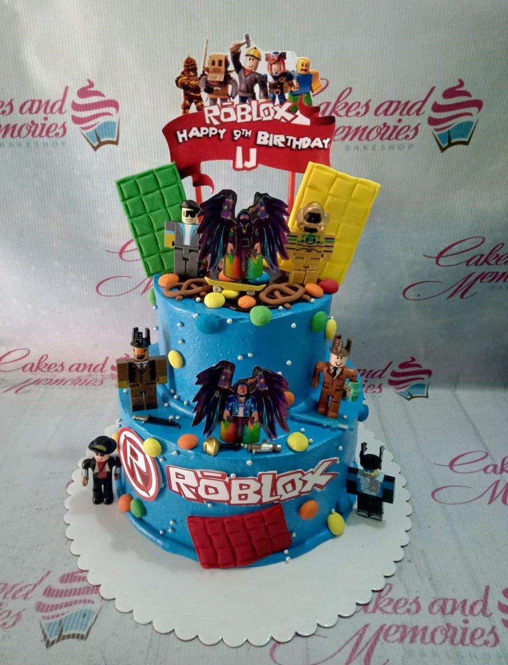 Blue 2-tier Roblox themed birthday cake with character printouts, chocolate bar decorations, and colorful gumpaste accents.