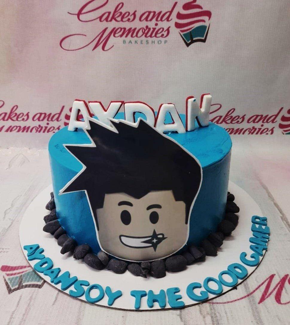 Blue Roblox themed birthday cake with a large character printout and black gumpaste stones around the base.