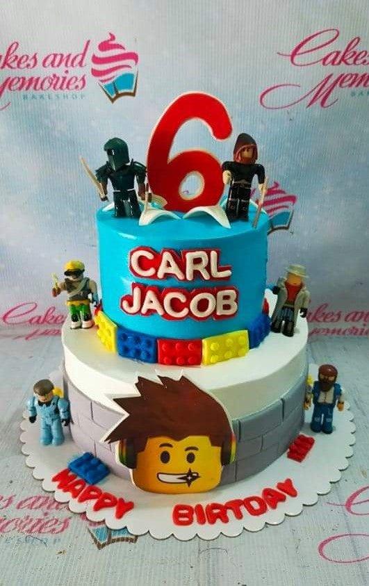 Two-tier blue and white Roblox themed birthday cake with Lego brick accents and character figurines.