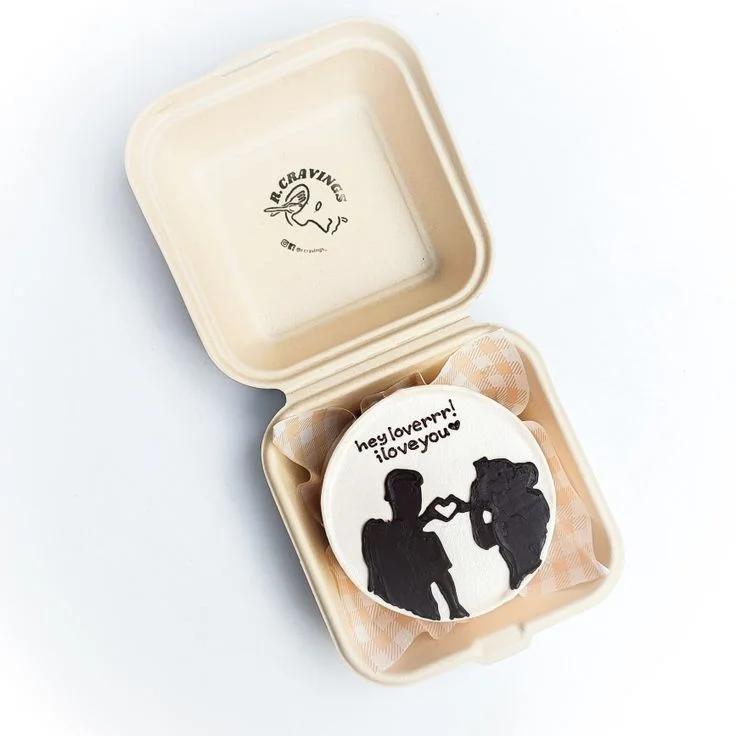 White bento cake with black icing silhouette of a couple making a heart shape and a romantic message in a bento box.