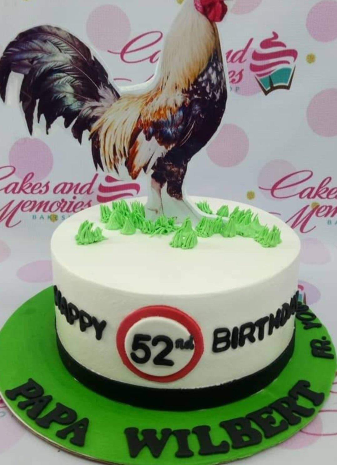White round cake with a large rooster printout topper, green icing grass, and a 52nd birthday message on the side.