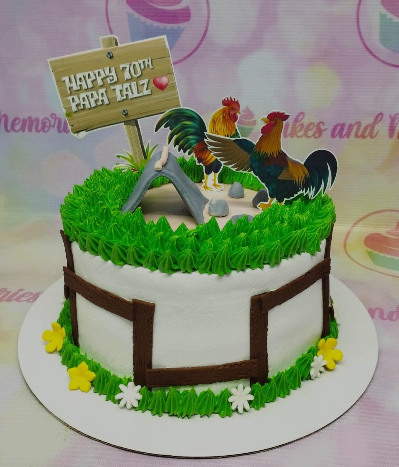White 1-tier rooster themed birthday cake with green icing grass, brown fondant fence, and rooster printouts for a 70th birthday.