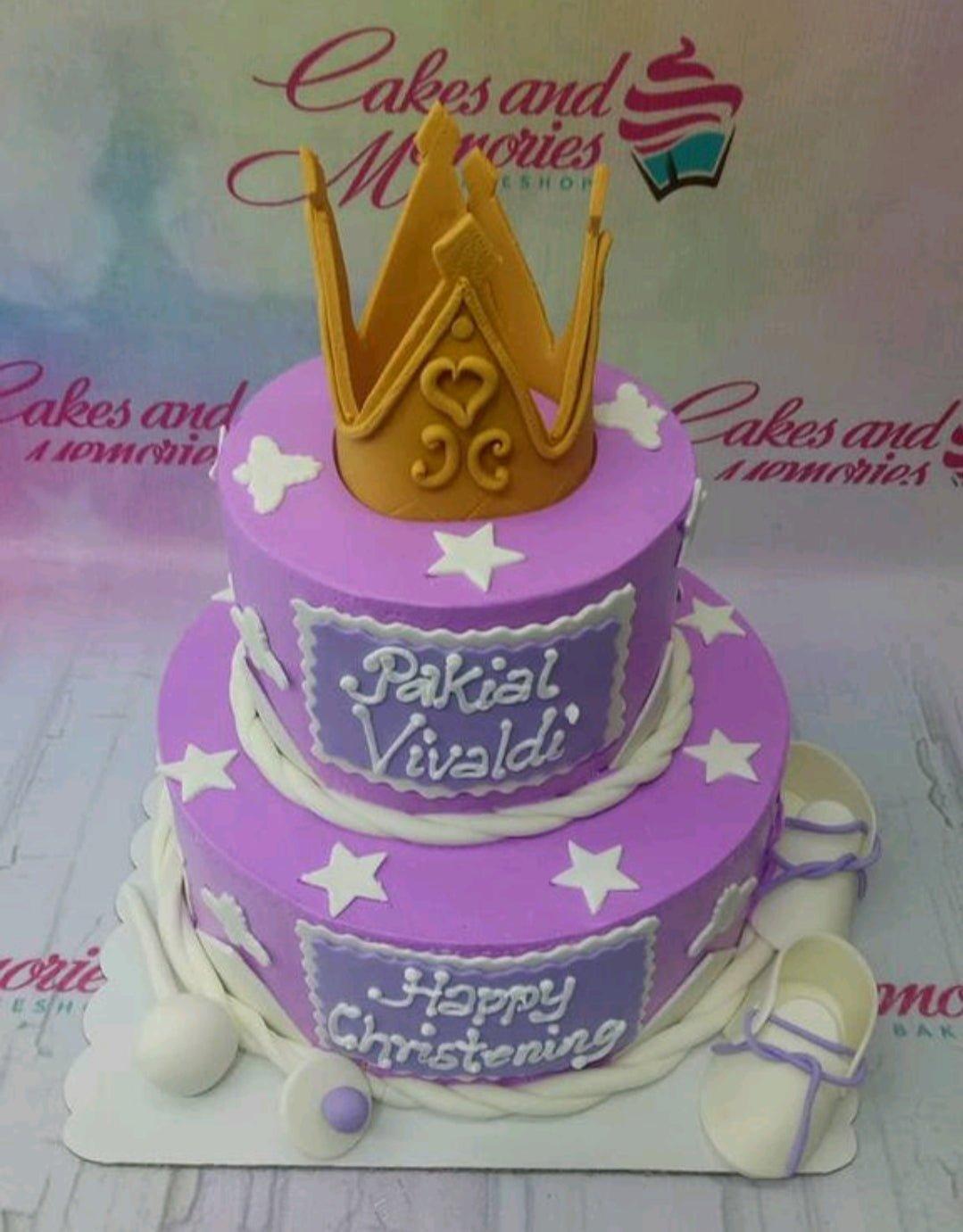 Two-tier purple fondant christening cake with a large gold crown topper, white stars, and baby shoes.