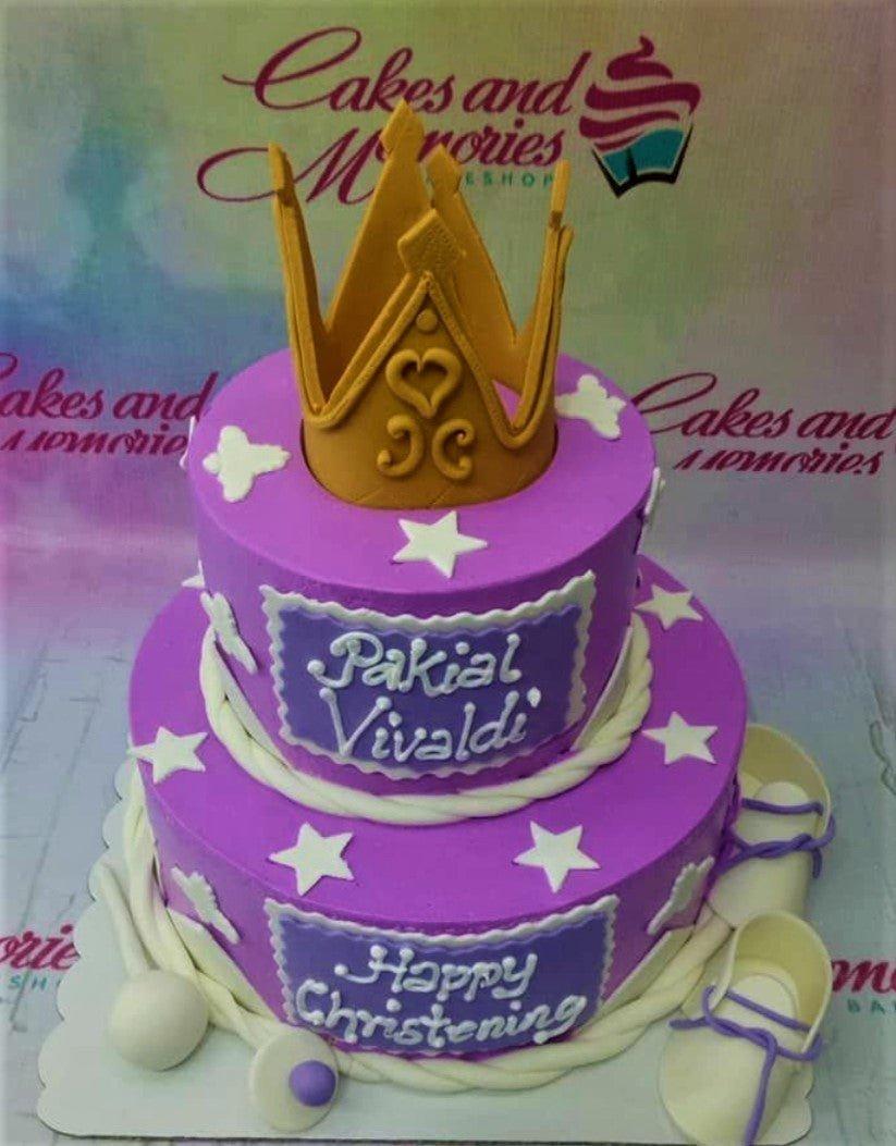 Two-tier purple fondant christening cake with a large gold crown topper, white stars, and baby shoes.