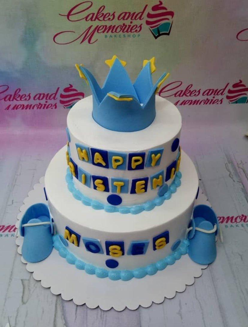 Two-tier white fondant cake with a blue crown topper, blue baby shoes, and blue and yellow gumpaste letter tiles.