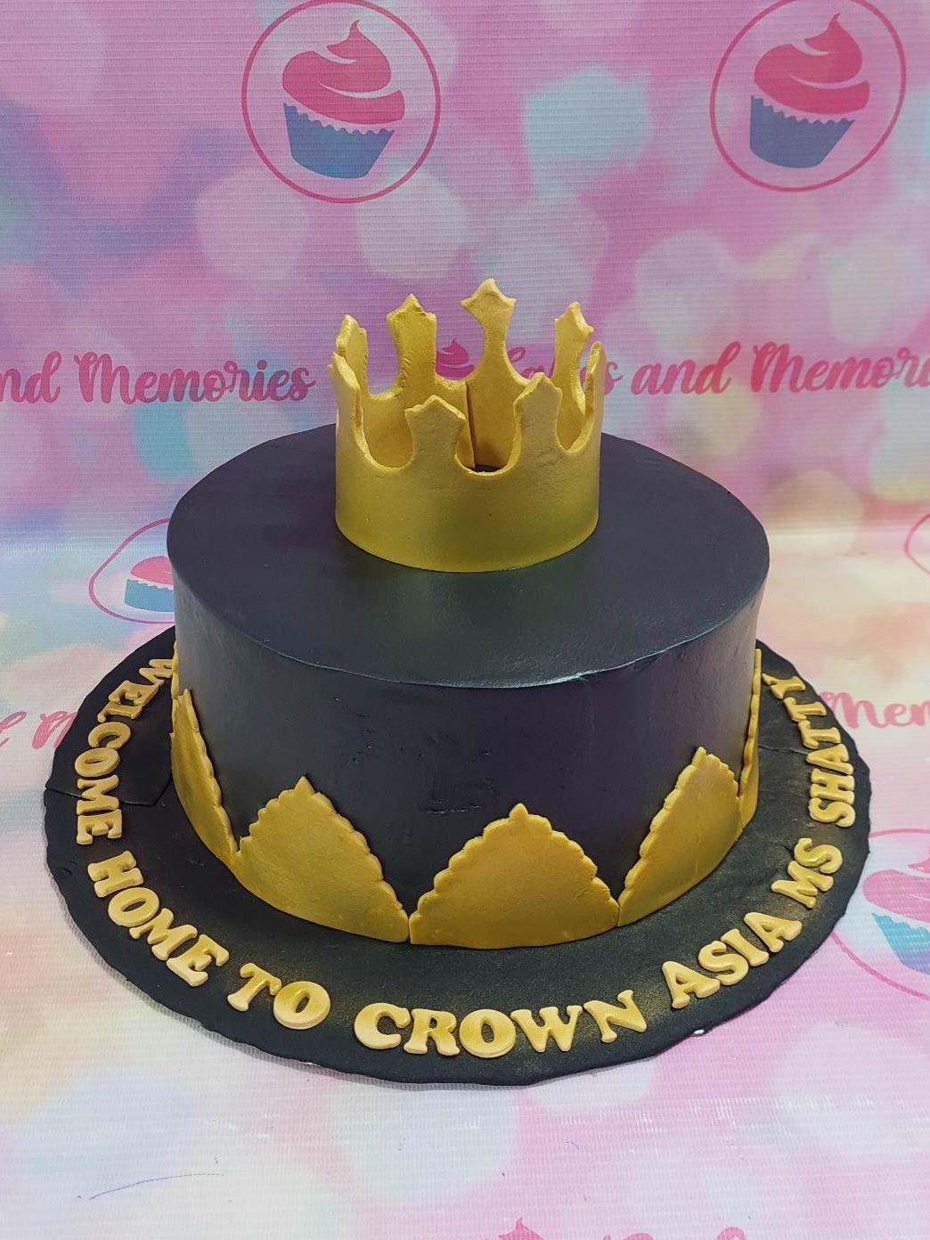 Black fondant cake with a large gold crown topper and gold leaf-like fondant accents around the base on a black board with gold lettering.