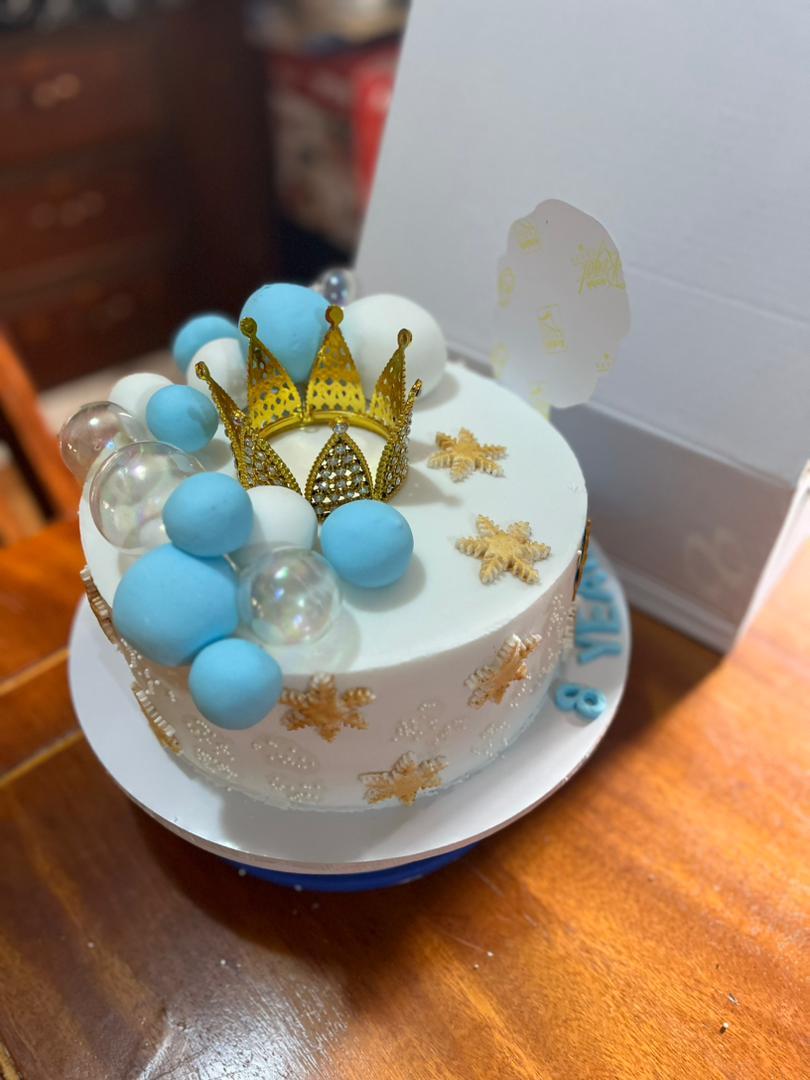 White round cake with a gold crown, blue and white fondant balls, and gold snowflakes.