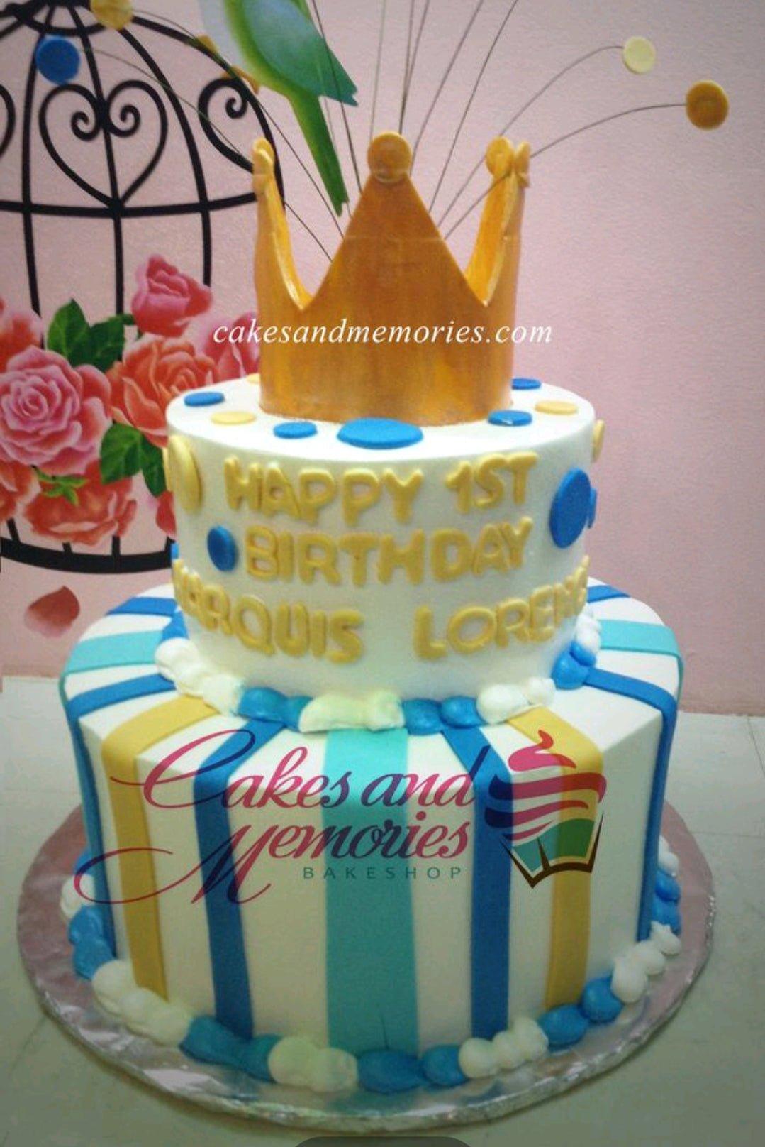 Two-tier white cake with blue and yellow vertical stripes, gold crown topper, and blue and yellow fondant dots.