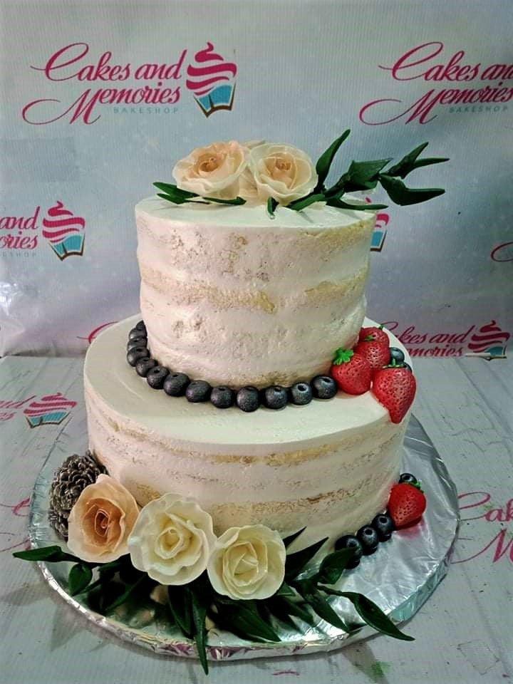Two-tier semi-naked cake decorated with fresh strawberries, blueberries, and cream-colored roses with green leaves on a silver board.